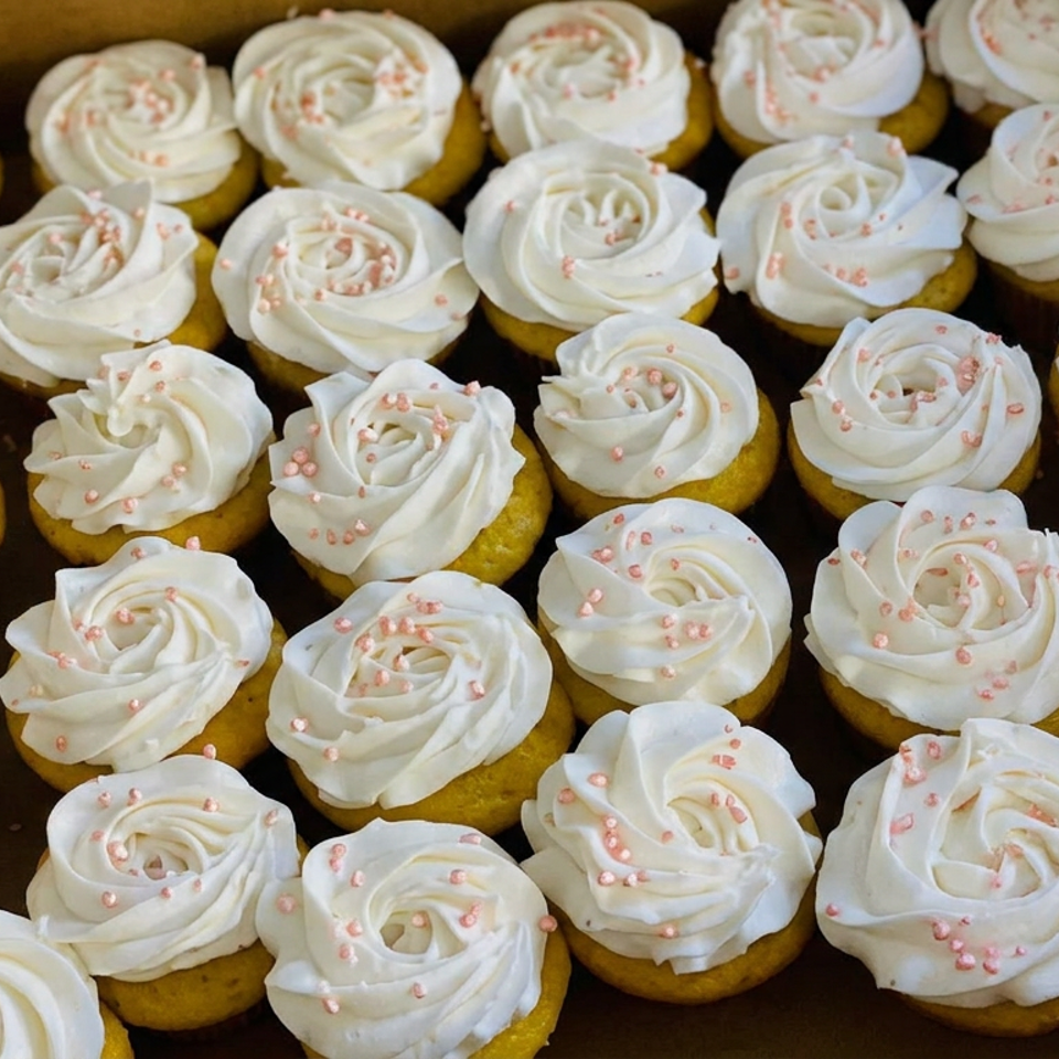 Box of yellow cupcakes topped with white swirled frosting and pink sprinkles.
