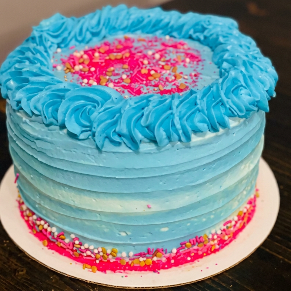 Blue frosted cake with piped rosette border, pink sprinkle center, and colorful sprinkles around the base on a wooden table.